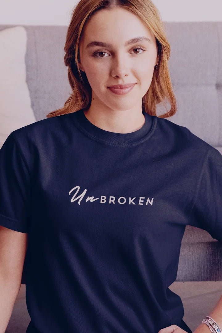 A woman wearing a navy blue crewneck t-shirt with the words 'Un BROKEN' printed in white script across the chest, posed in front of a gray couch.