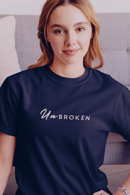 A woman wearing a navy blue crewneck t-shirt with the words 'Un BROKEN' printed in white script across the chest, posed in front of a gray couch.