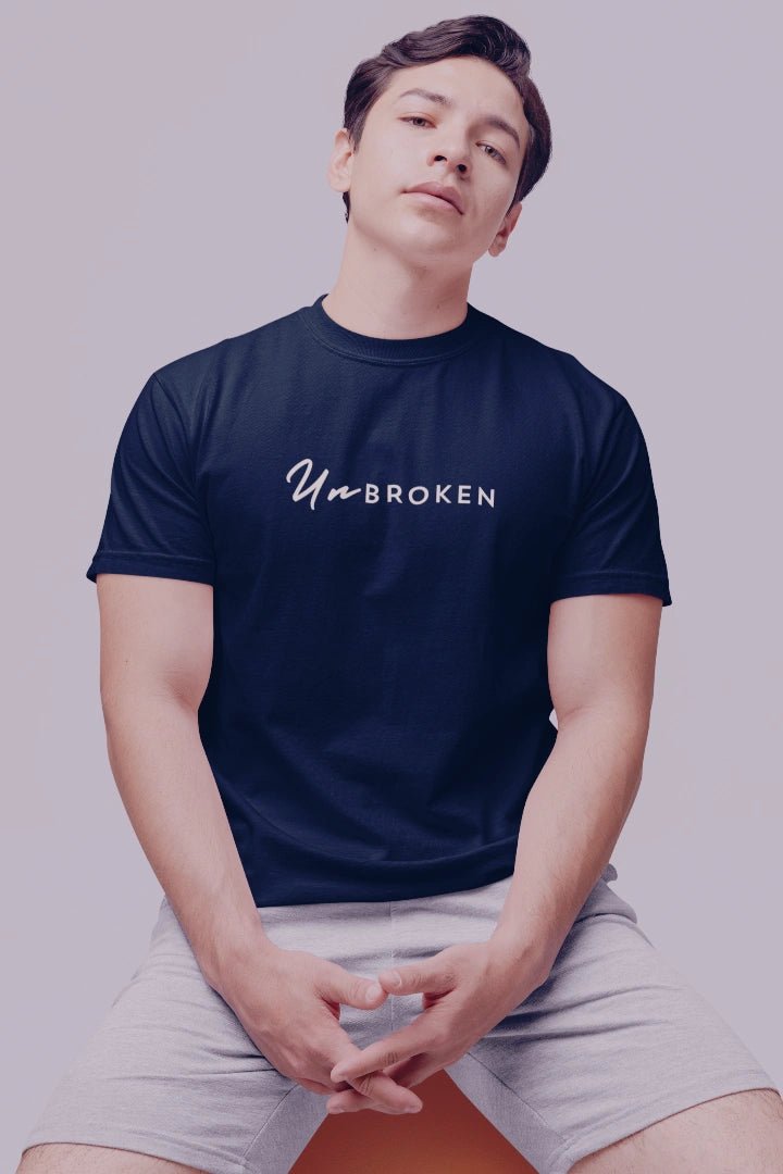 A man wearing a navy blue crewneck t-shirt with the text 'UnBROKEN' printed in white across the chest, paired with light gray shorts, seated against a solid light gray background.