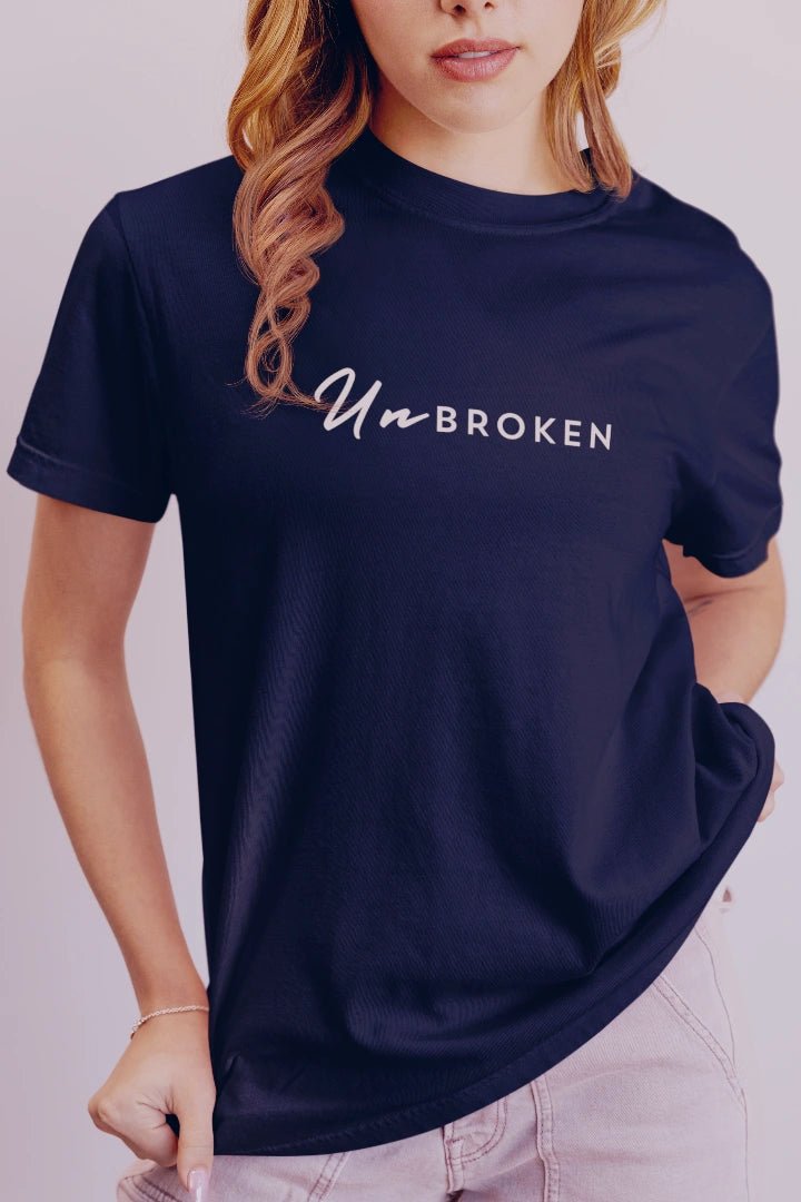 A woman wearing a navy blue short-sleeve t-shirt with the text 'UnbROKEN' printed in white across the chest, paired with light pink jeans against a light gray studio background.