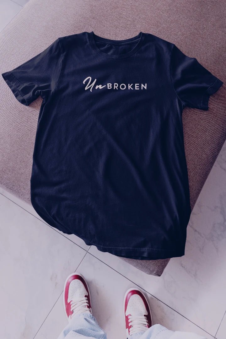 A navy blue t-shirt with 'UnBROKEN' printed in white across the chest, laid flat on a textured brown surface with a person's feet in red and white sneakers visible at the bottom.