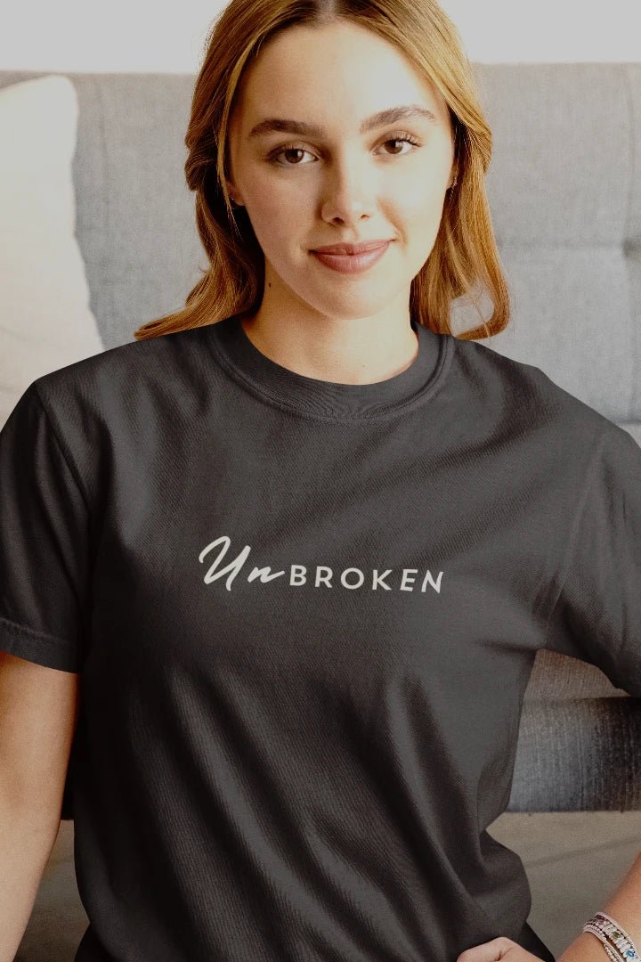 A woman wearing a steel grey t-shirt with the words 'UnBROKEN' printed in white across the chest, posed in front of a gray couch.