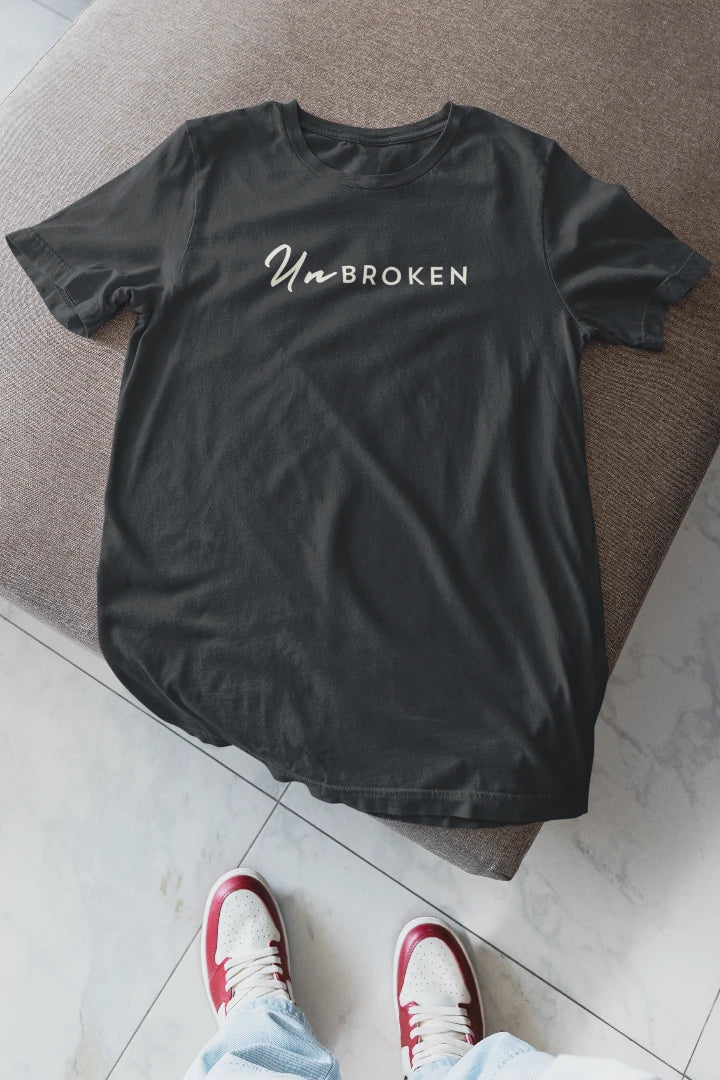 A steel grey short-sleeve t-shirt with 'UnBROKEN' printed in white across the chest, laid flat on a brown upholstered bench with a person's legs and red-and-white sneakers visible at the bottom.