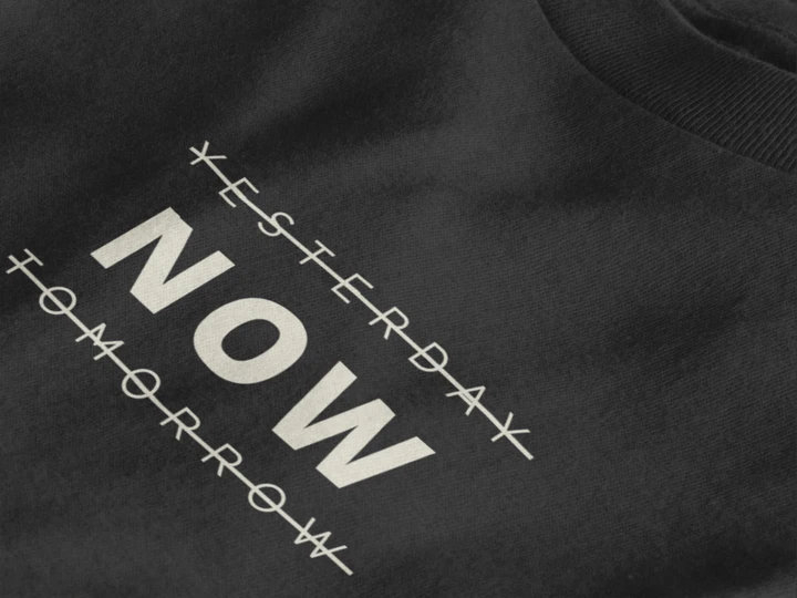 Close-up of a jet black crewneck t-shirt featuring a black graphic print that reads 'NOW' crossed out with lines above and below it, with 'YESTERDAY' and 'TOMORROW' 