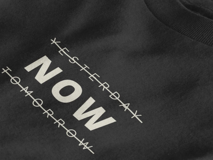 Close-up of a jet black crewneck t-shirt featuring a black graphic print that reads 'NOW' crossed out with lines above and below it, with 'YESTERDAY' and 'TOMORROW' 
