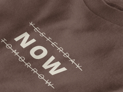 Close-up of a dark mocha crewneck t-shirt featuring a black graphic print that reads 'NOW' crossed out with lines above and below it, with 'YESTERDAY' and 'TOMORROW' 