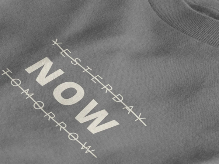 Close-up of a heather charcoal crewneck t-shirt featuring a black graphic print that reads 'NOW' crossed out with lines above and below it, with 'YESTERDAY' and 'TOMORROW' 