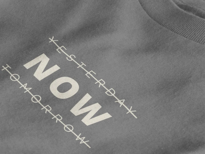 Close-up of a heather charcoal crewneck t-shirt featuring a black graphic print that reads 'NOW' crossed out with lines above and below it, with 'YESTERDAY' and 'TOMORROW' 