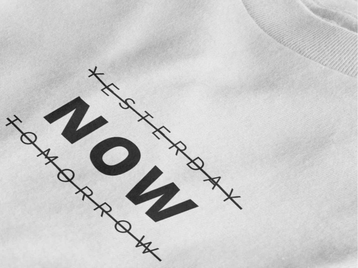 Close-up of a pure white crewneck t-shirt featuring a black graphic print that reads 'NOW' crossed out with lines above and below it, with 'YESTERDAY' and 'TOMORROW' 