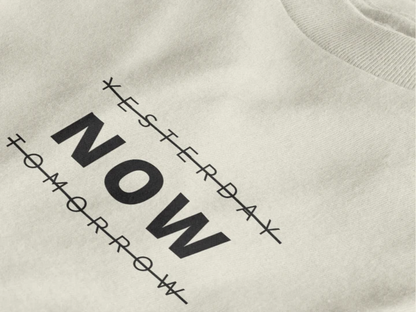 Close-up of a off white crewneck t-shirt featuring a black graphic print that reads 'NOW' crossed out with lines above and below it, with 'YESTERDAY' and 'TOMORROW' 