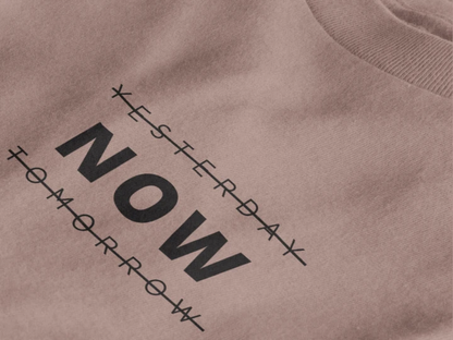 Close-up of a taupe crewneck t-shirt featuring a black graphic print that reads 'NOW' crossed out with lines above and below it, with 'YESTERDAY' and 'TOMORROW' 
