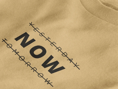 Close-up of a warm sand crewneck t-shirt featuring a black graphic print that reads 'NOW' crossed out with lines above and below it, with 'YESTERDAY' and 'TOMORROW' 
