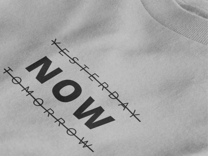 Close-up of a heather grey crewneck t-shirt featuring a black graphic print that reads 'NOW' crossed out with lines above and below it, with 'YESTERDAY' and 'TOMORROW' 